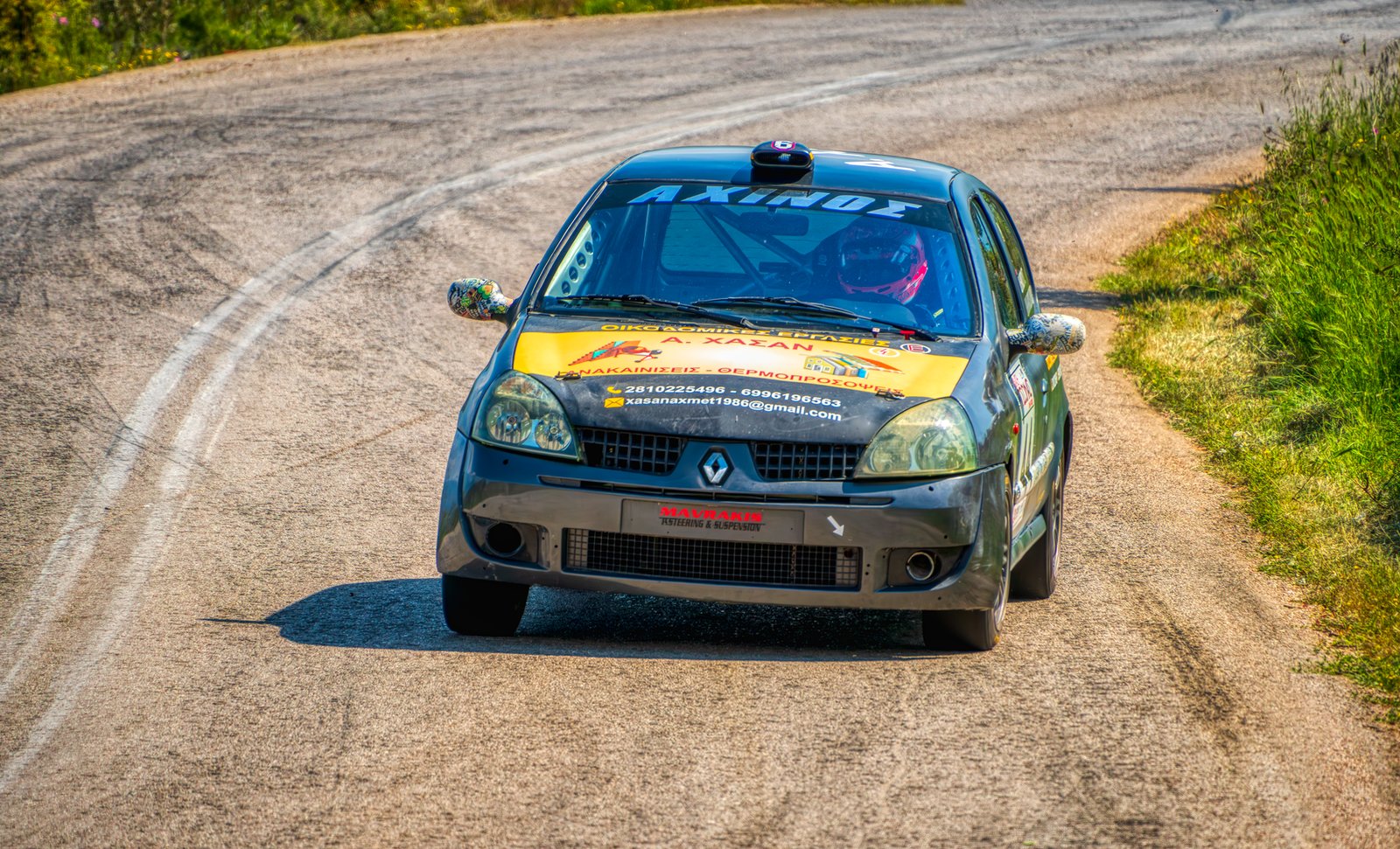 Rally car with vibrant decals races on a sunny, winding road in Greece.
