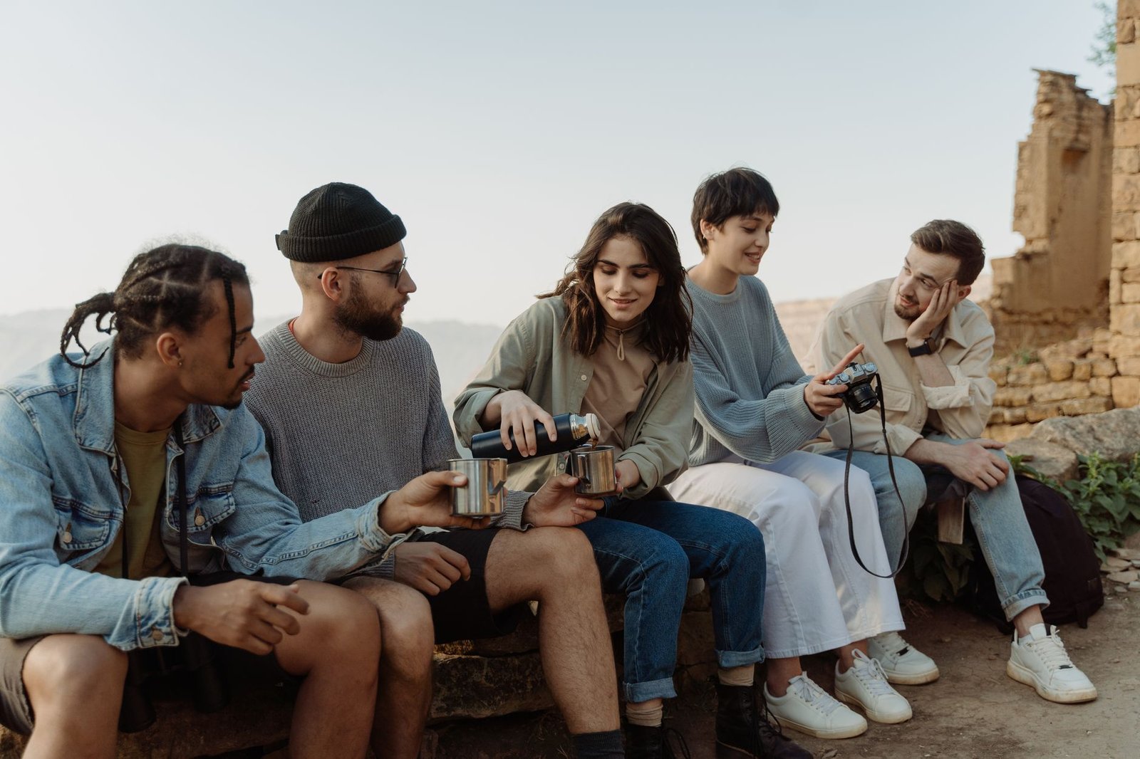 A diverse group of friends enjoy hiking together, sitting and sharing drinks outdoors on a sunny day.