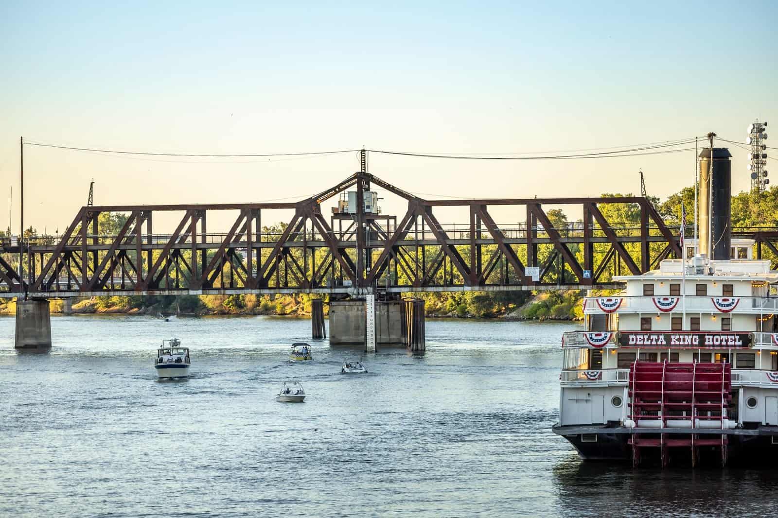 View of the Delta King Hotel and historic bridge over a river with small boats passing by.