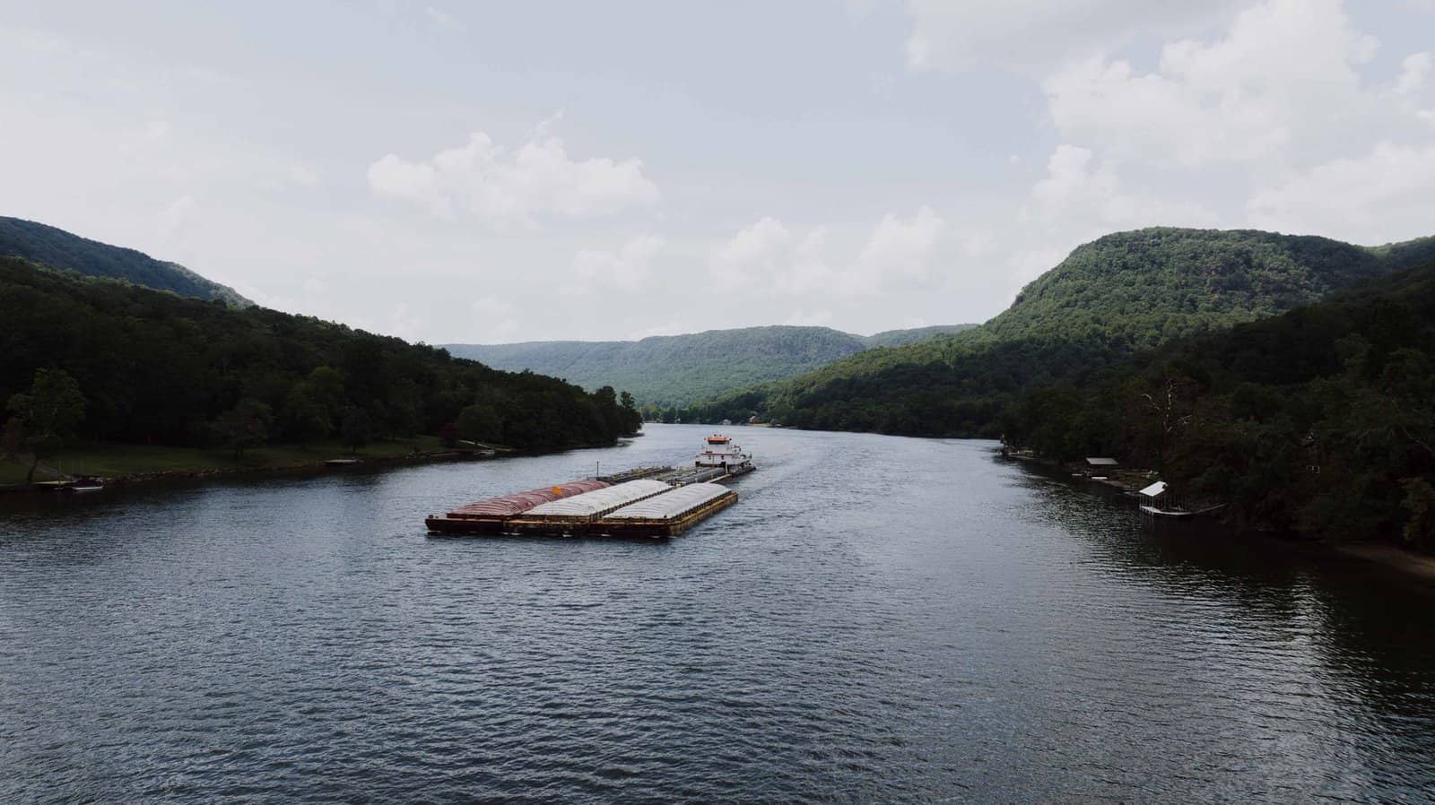 A serene aerial shot of a barge navigating the Tennessee River surrounded by lush hills and summer skies.