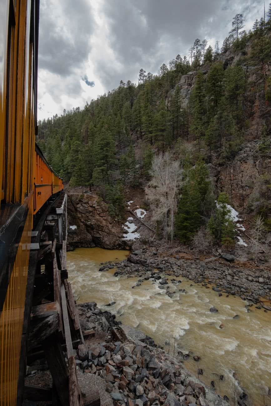 A train crossing a wooden bridge over a river in a lush mountain landscape during fall.