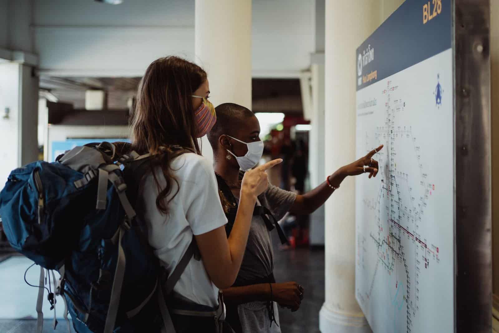 Two women with backpacks navigate a transit map, wearing face masks.