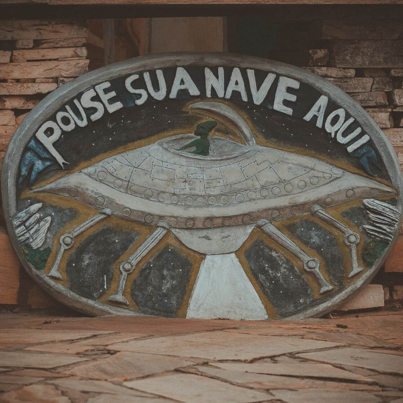 Painted oval stone sign depicting a UFO with Portuguese text.