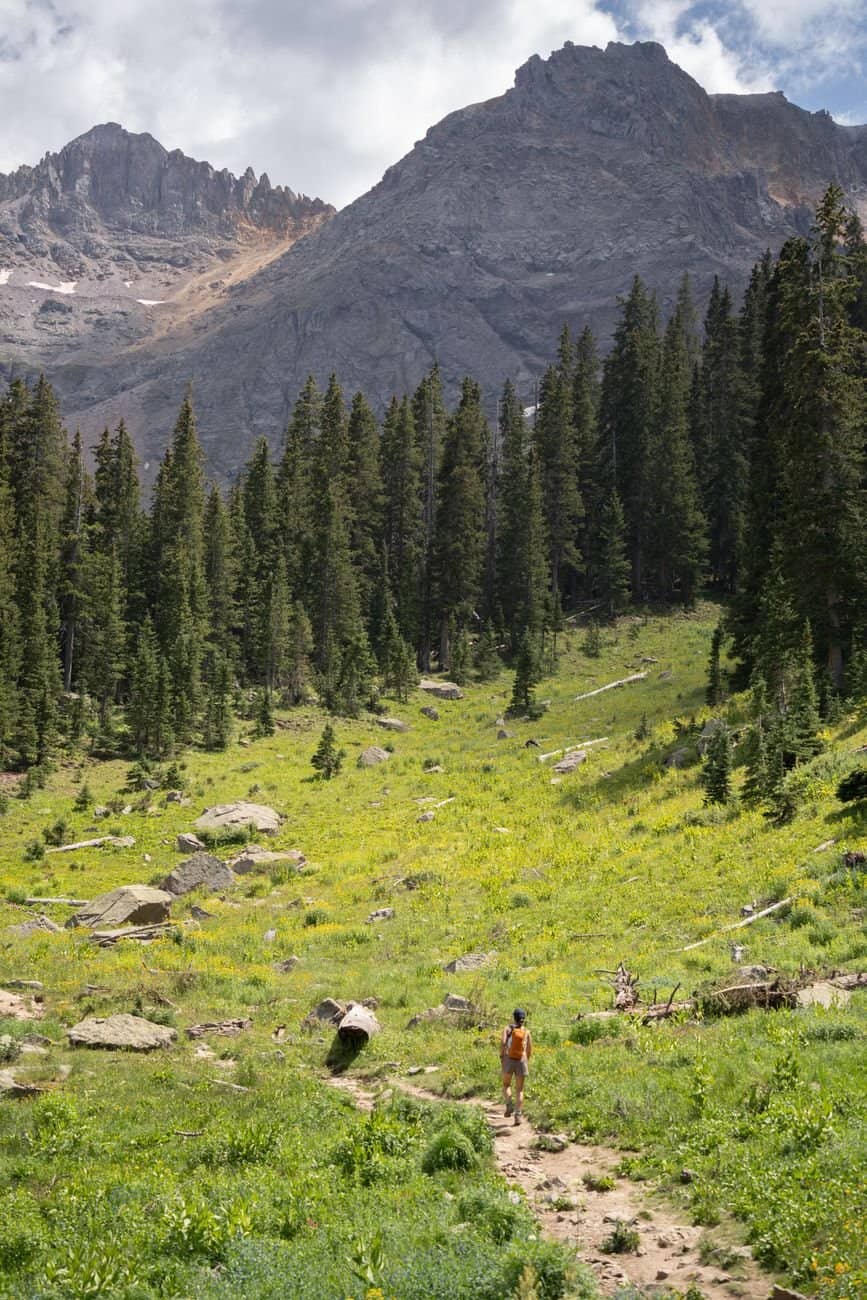 A hiker explores a lush mountain trail surrounded by evergreen forests under a clear summer sky.