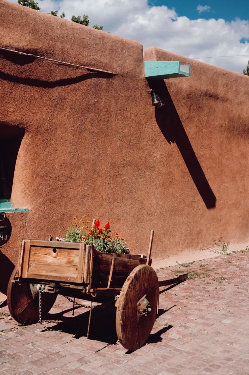 Sunlit rustic wooden cart with flowers beside an orange adobe wall under a blue sky.