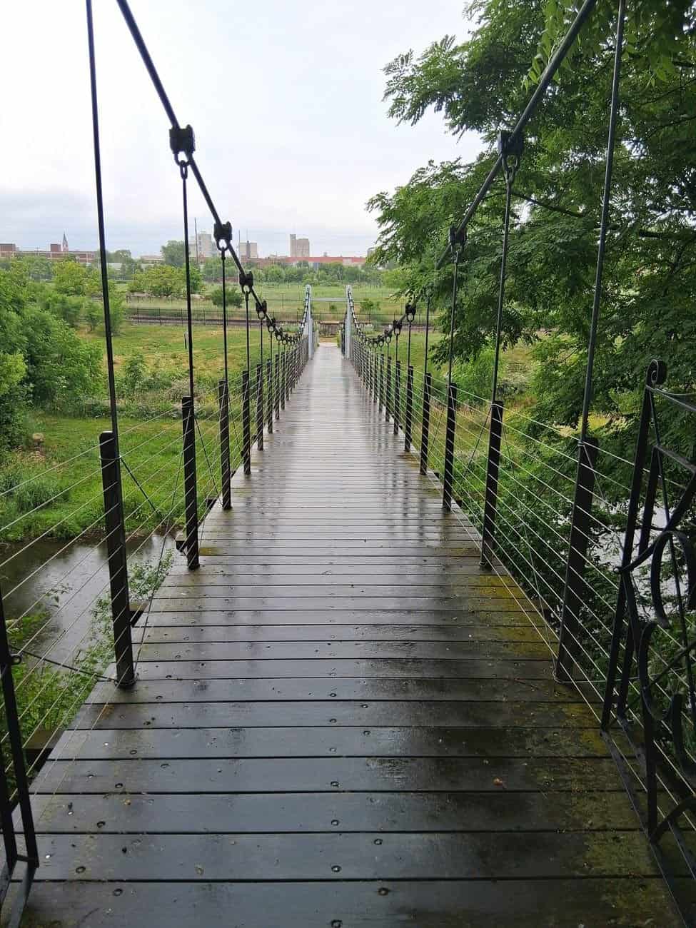 A suspension footbridge extends over vibrant green foliage, under a lightly overcast sky.