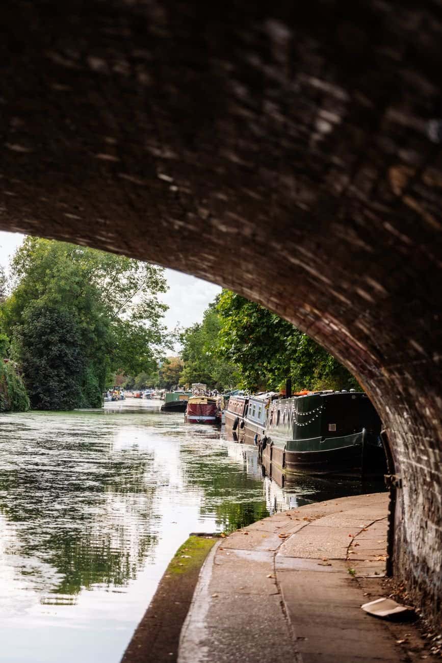 Serene canal view with houseboats under a historic brick arch in London.