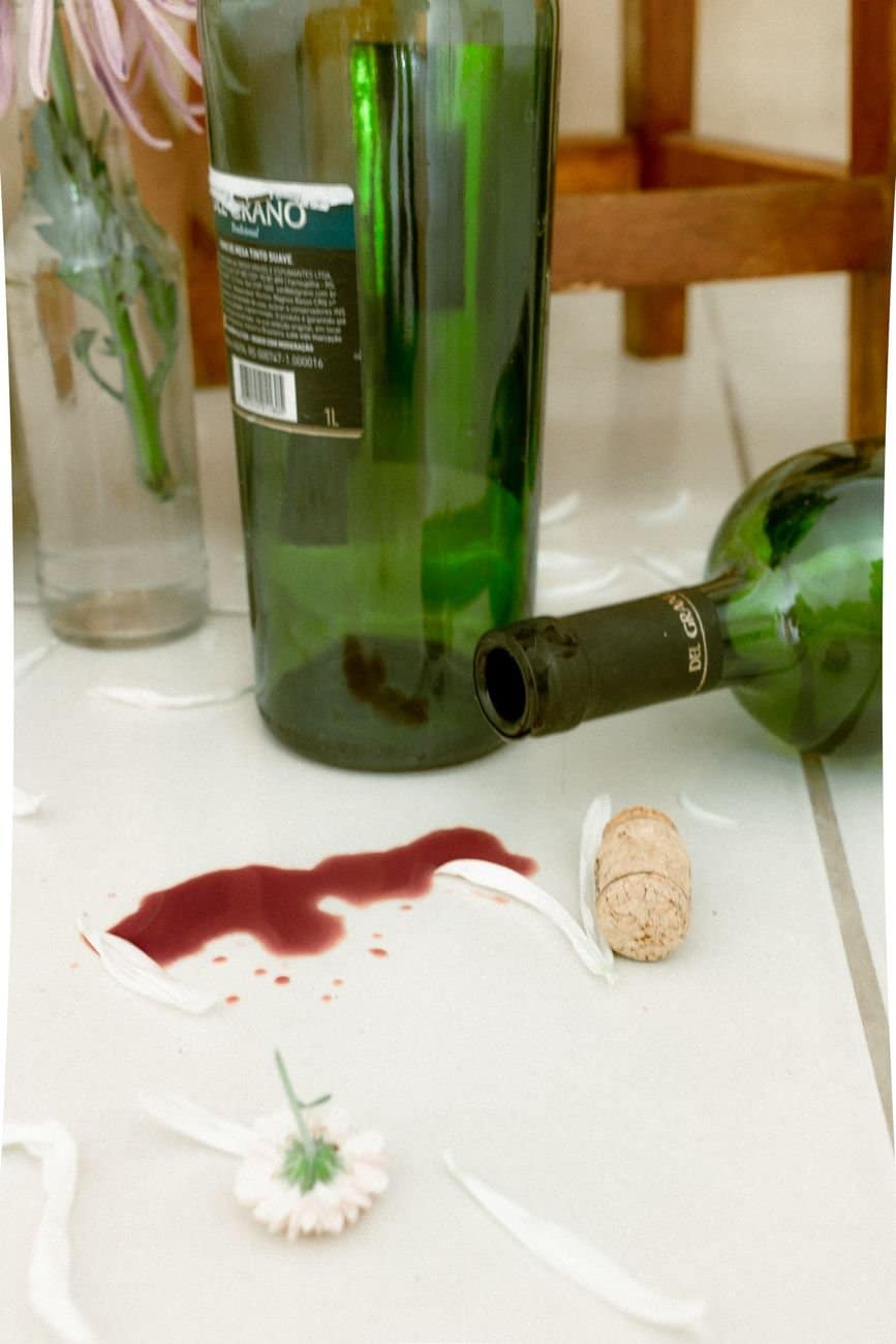 A still life of red wine spill, glass bottles, and flowers on a ceramic floor.