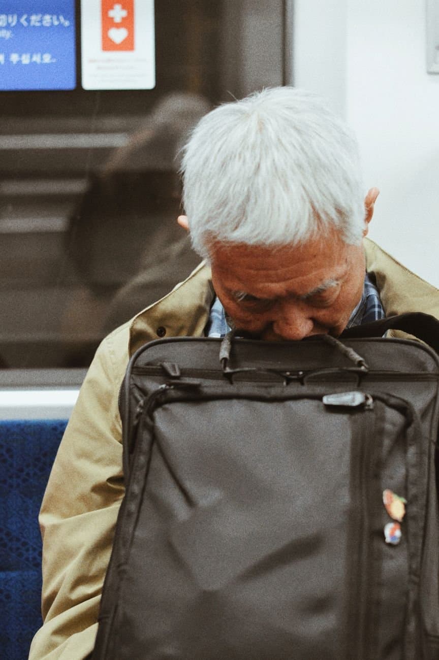 An elderly man asleep on a subway train, resting his head on a backpack.