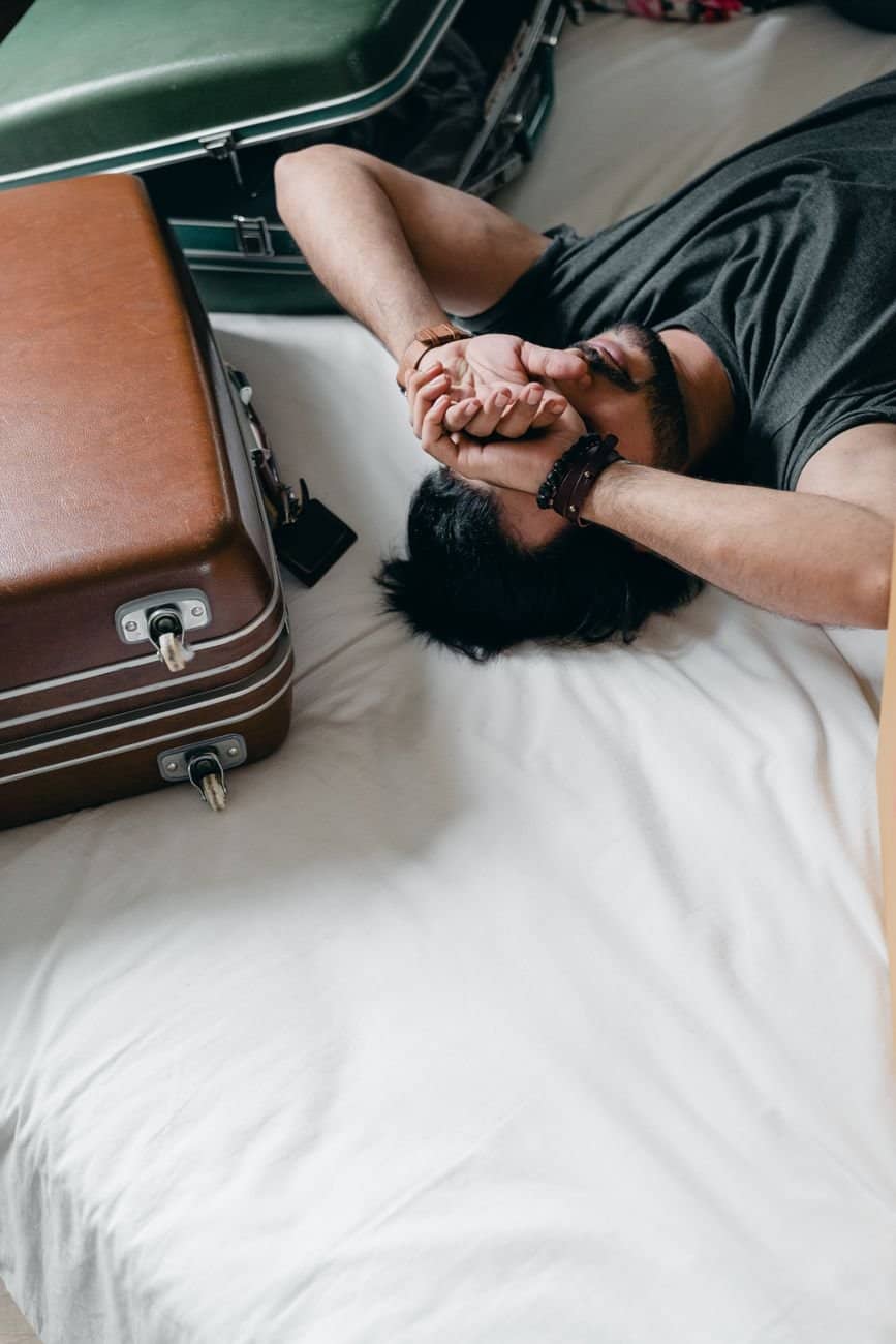 From above young bearded male wearing gray t shirt resting on bed near suitcases and covering eyes with arms while moving into new apartment