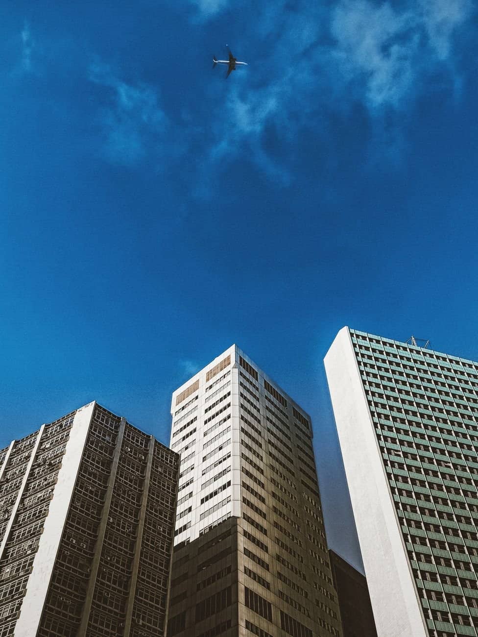 From below of contemporary multistory city building exteriors with shadow under aircraft flying in cloudy sky on sunny day