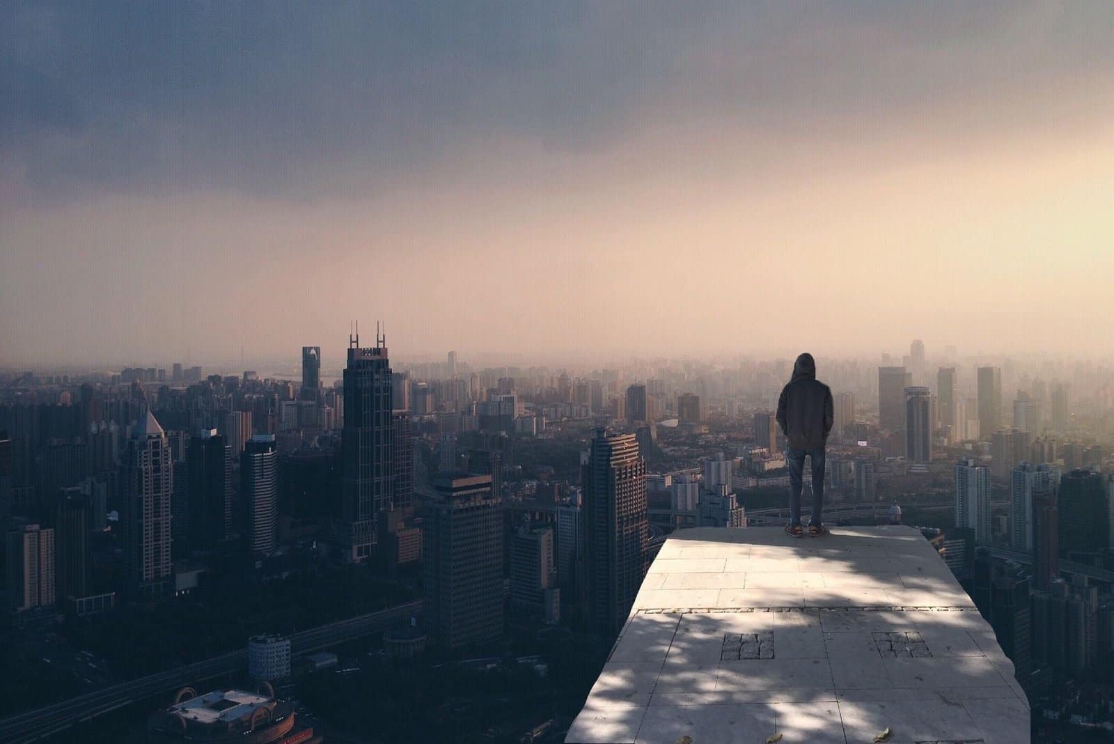 A person stands on a rooftop, gazing over a foggy city skyline at dusk.