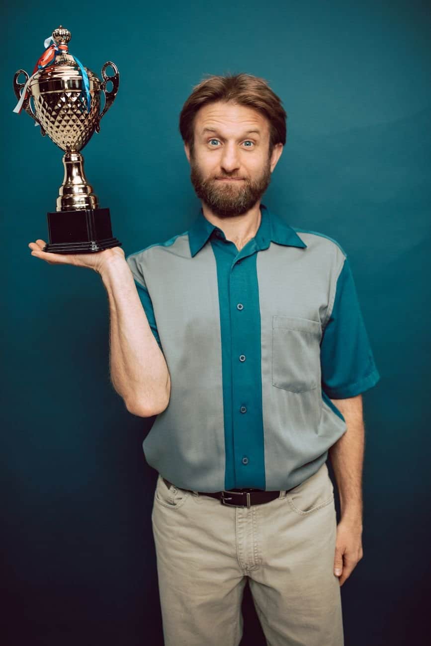 A bearded man confidently holds a trophy in a studio setting, showcasing victory and accomplishment.