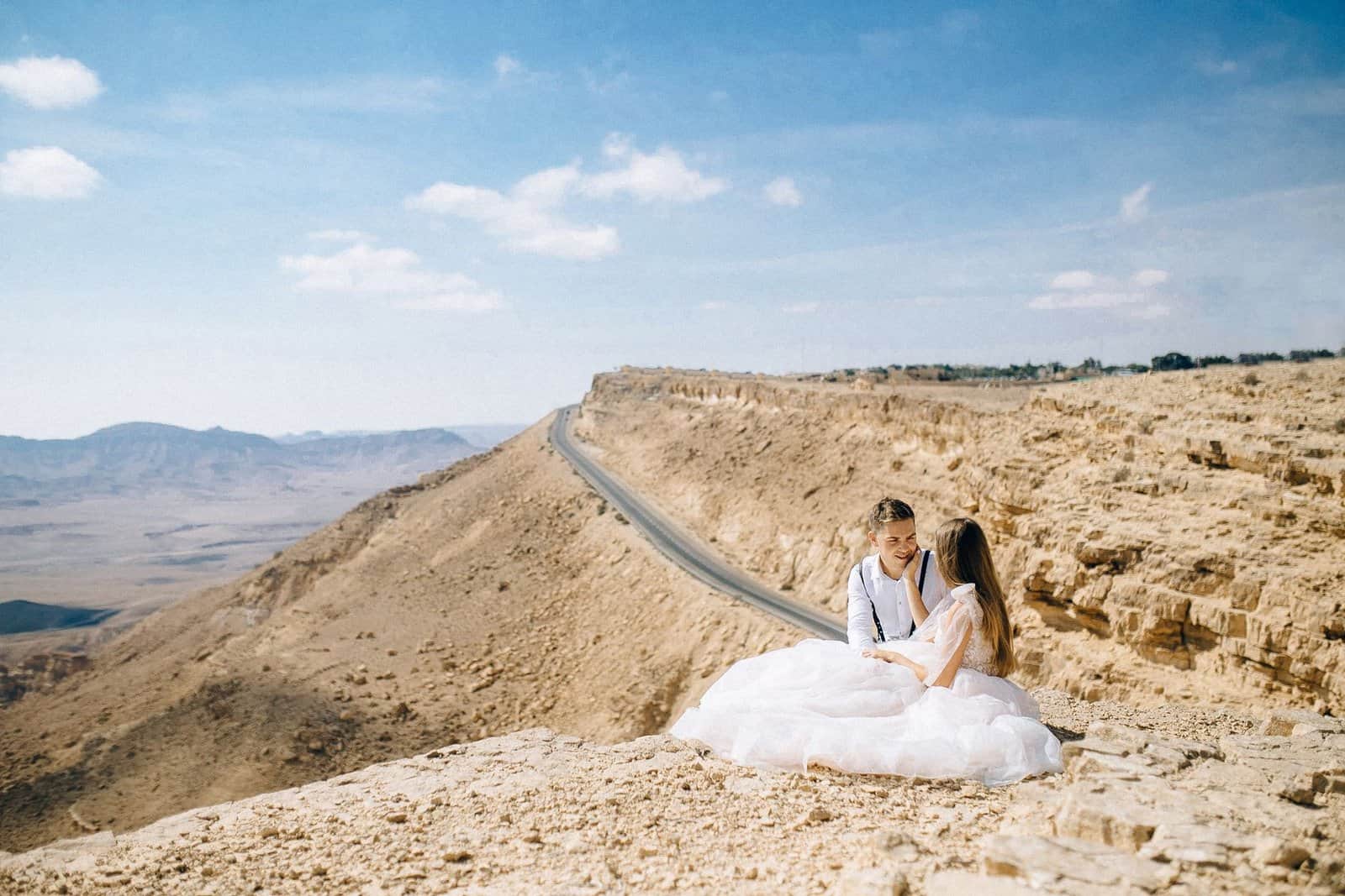 Couple in wedding attire sitting on a cliff, embracing the vast desert view.