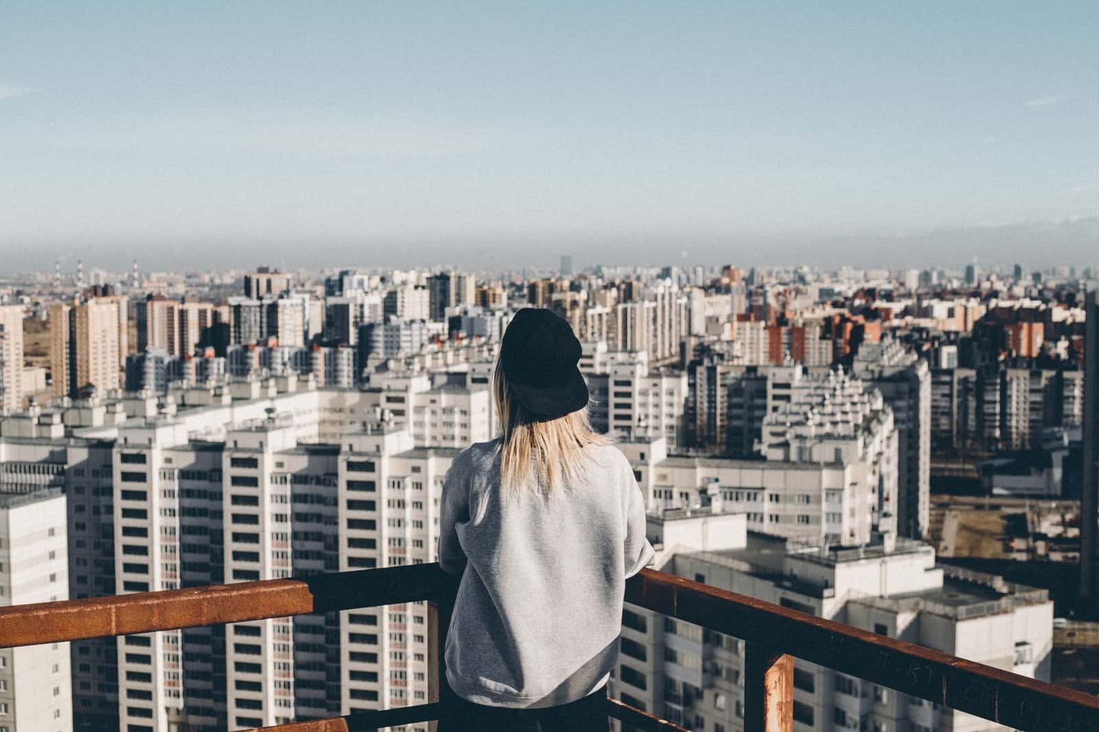 A person in a black cap and sweatshirt admires a vast cityscape from a rooftop perspective.