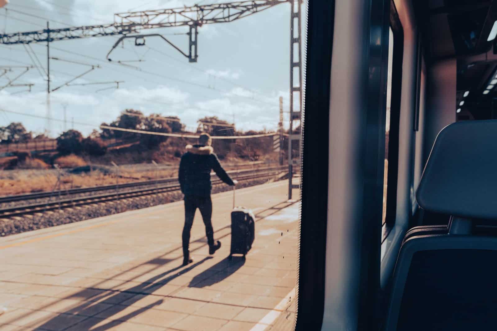 A man walks on a train platform with luggage, viewed from a train window.