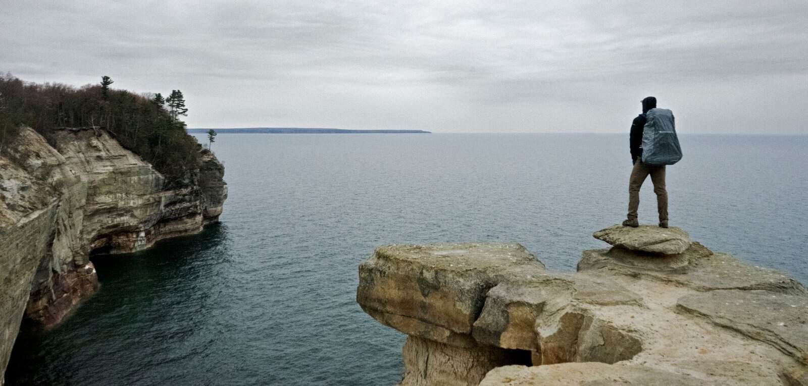 A man with a backpack stands on a rocky cliff at Pictured Rocks, Michigan, gazing over Lake Superior.