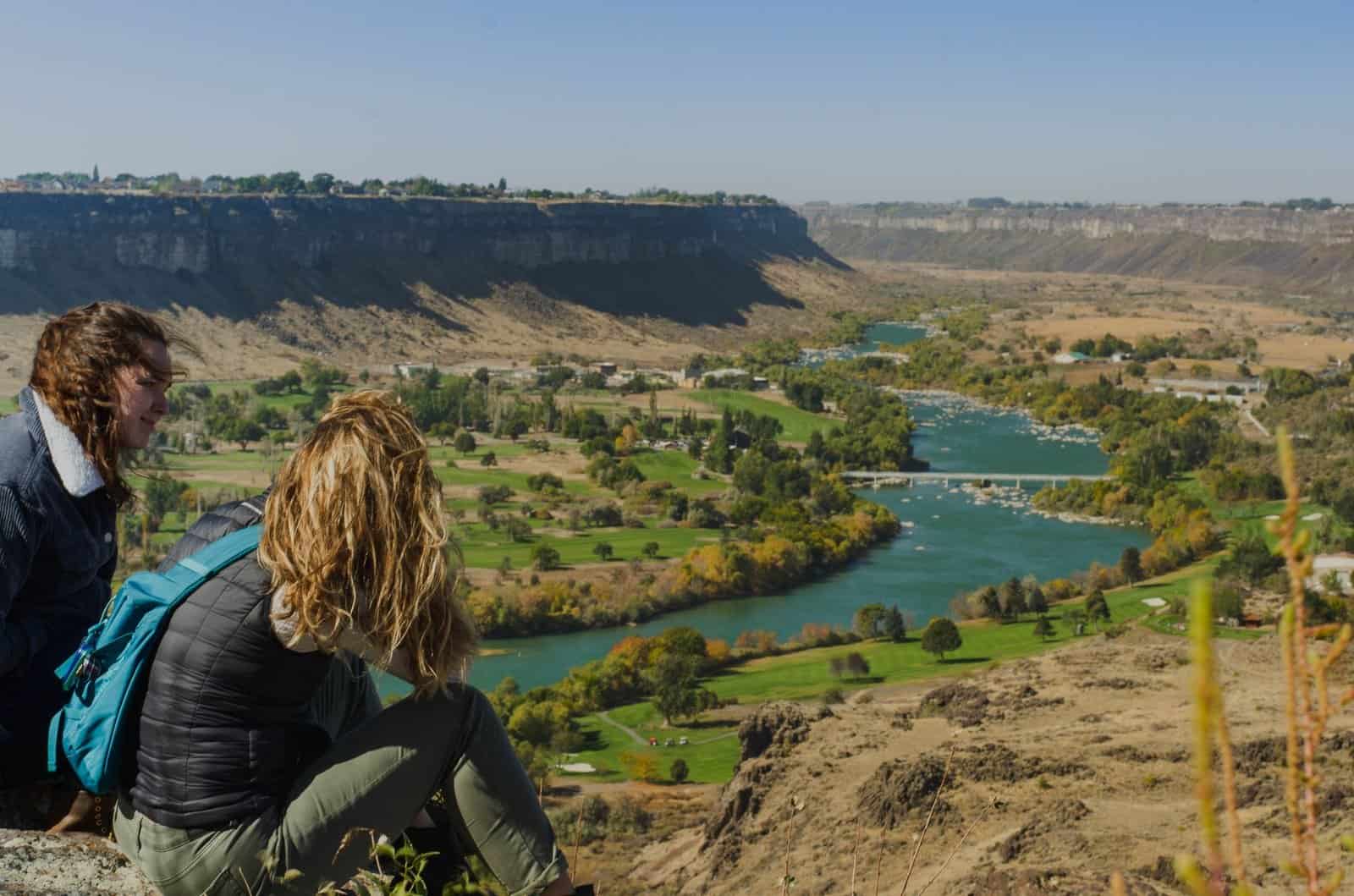 Two women sitting on a cliff enjoying a scenic view of a river valley with lush greenery.