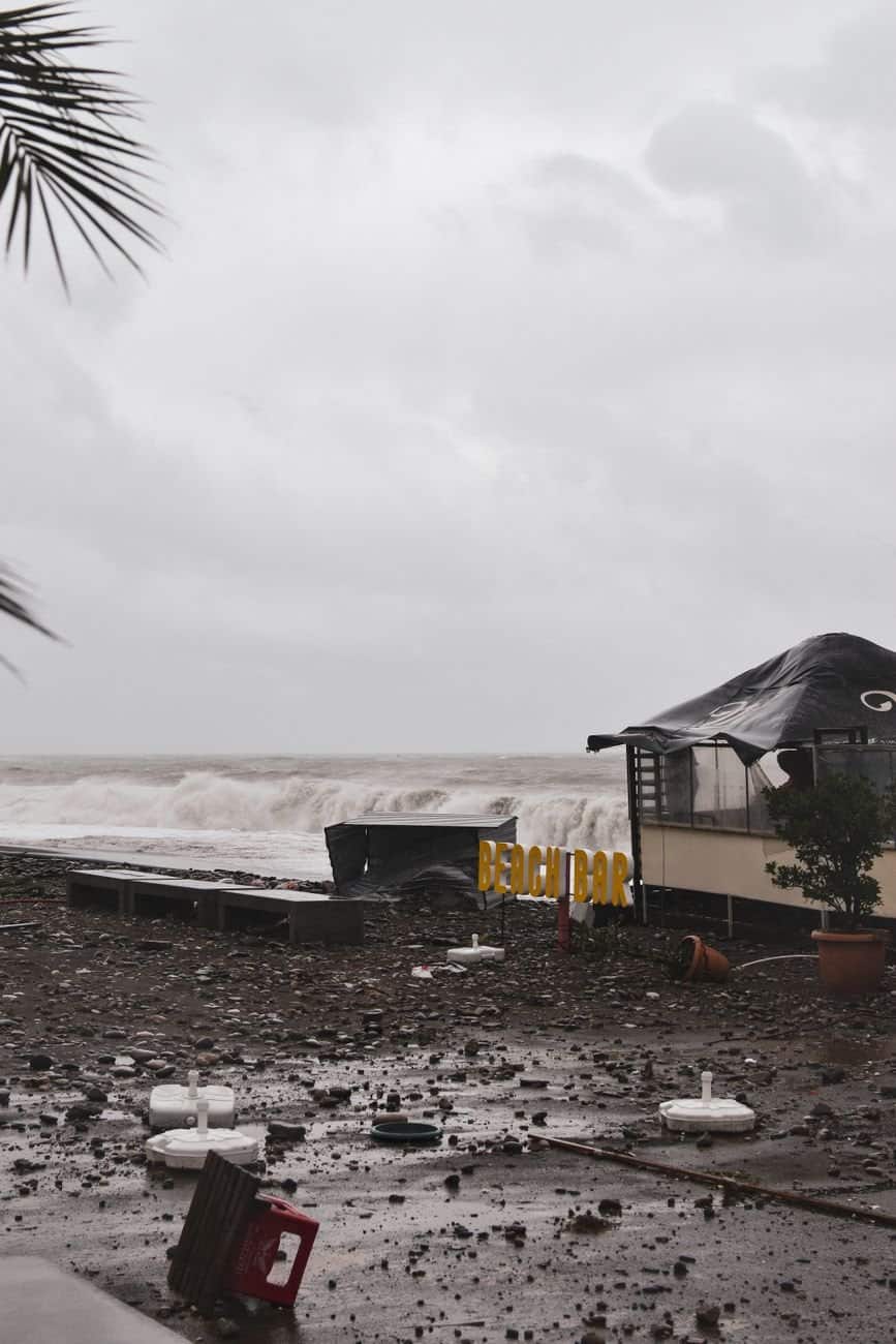 A coastal beach bar damaged by a recent storm with high waves, debris, and cloudy skies.