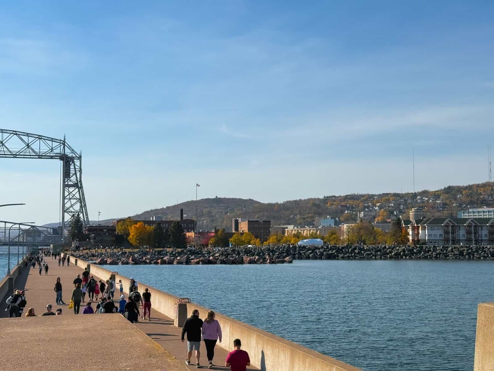 People enjoy a sunny day by the Duluth Aerial Lift Bridge with vibrant fall foliage.