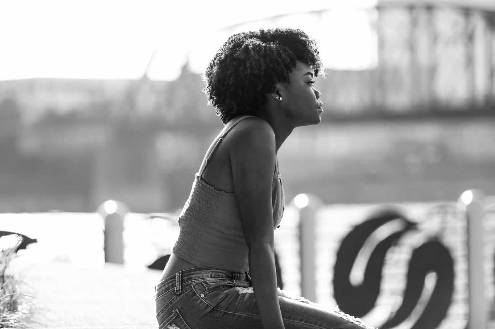 Black and white portrait of a woman with natural hair sitting by a river, reflecting deep in thought.