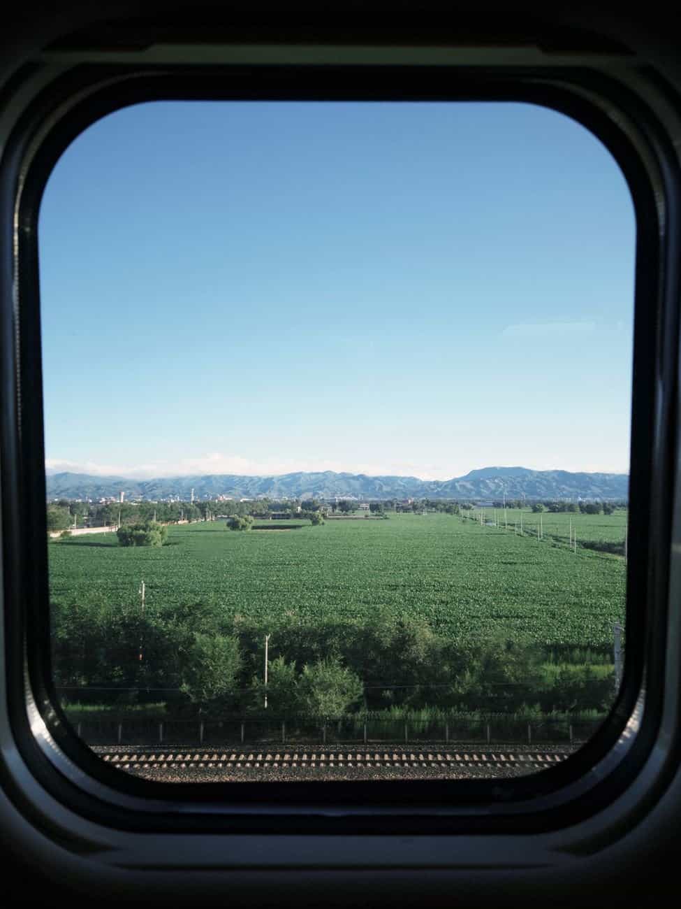 View of lush green fields and mountains from a train window on a sunny day.