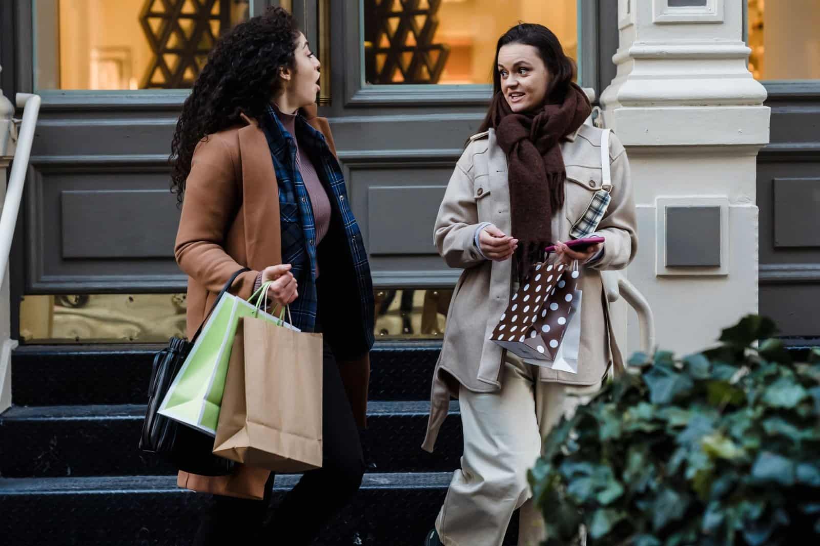 Young stylish lady discussing news with surprised ethnic female friend while walking together on city street after shopping
