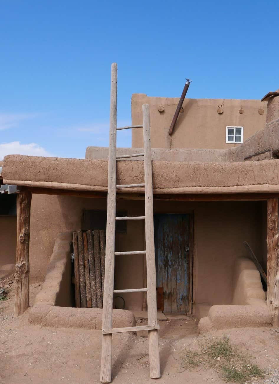 Rustic adobe building with a wooden ladder in Taos, New Mexico under a clear blue sky.