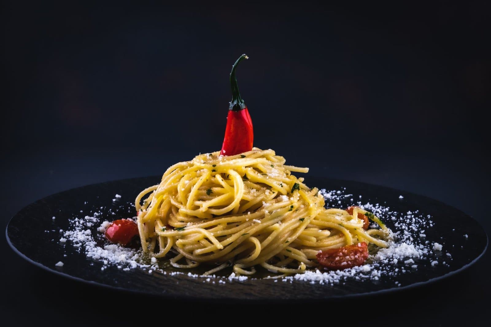 A beautifully styled plate of spaghetti with chili pepper on a black dish, garnished with cheese.