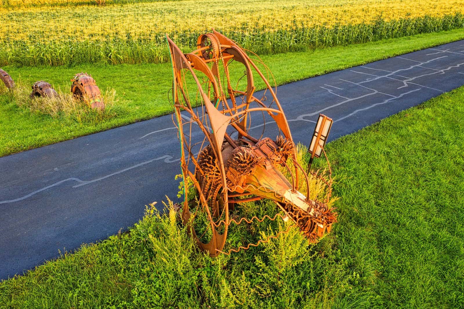 A unique rusty sculpture on a rural road in Minnesota near lush green fields.